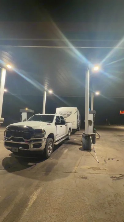 A white Ram truck towing an RV rental at a lit gas station at night. A white cane rests by the pump, preparing for the road.. Crossroads RV Rezerve 2015