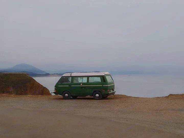 Green VW camper van RV rental on a cliff overlooking a misty ocean. Perfect for a scenic road trip adventure.. Volkswagen Vanagon 1984