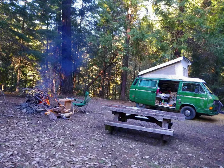 Forest campsite with a crackling campfire, a green RV rental van (pop-top open), and a wooden picnic table. Van life adventure awaits.. Volkswagen Vanagon 1984