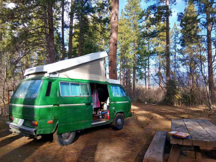 Green VW Westfalia camper, perfect as an RV rental, parked among tall pines at a peaceful campsite with a picnic table.. Volkswagen Vanagon 1984