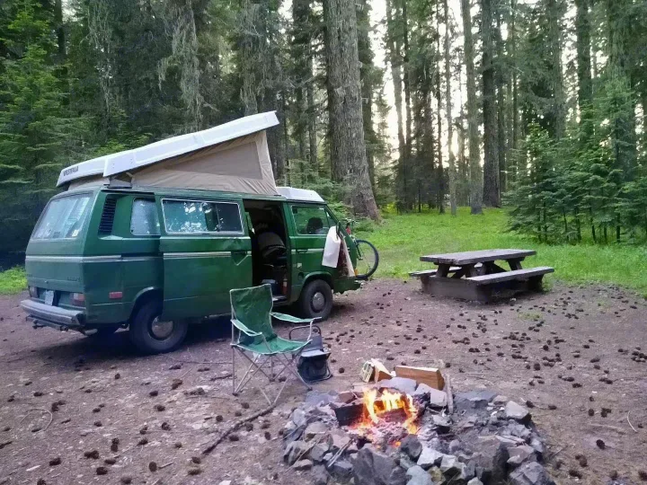 Green Westfalia camper van with pop-top, campfire, picnic table, and chair at a forest camp. Great for an RV rental.. Volkswagen Vanagon 1984