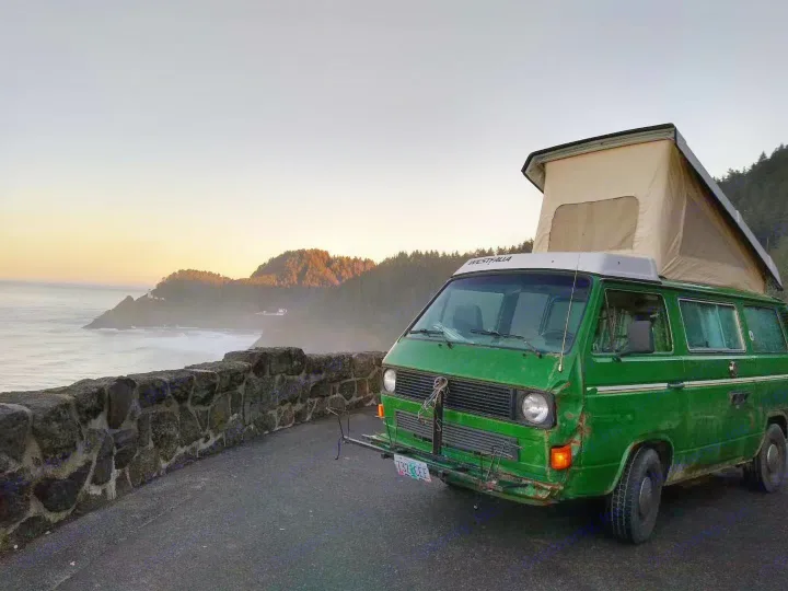A vibrant green Westfalia pop-top camper van is parked on a coastal road, overlooking a breathtaking sunset over the ocean. This classic RV rental is ready for adventure.. Volkswagen Vanagon 1984