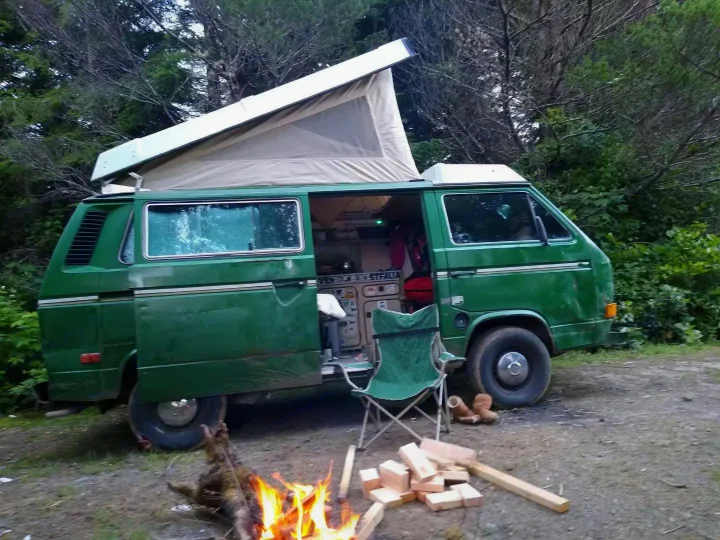 Experience camping: Green VW Westfalia pop-top camper van, an ideal rv rental, set up by a campfire in the woods.. Volkswagen Vanagon 1984