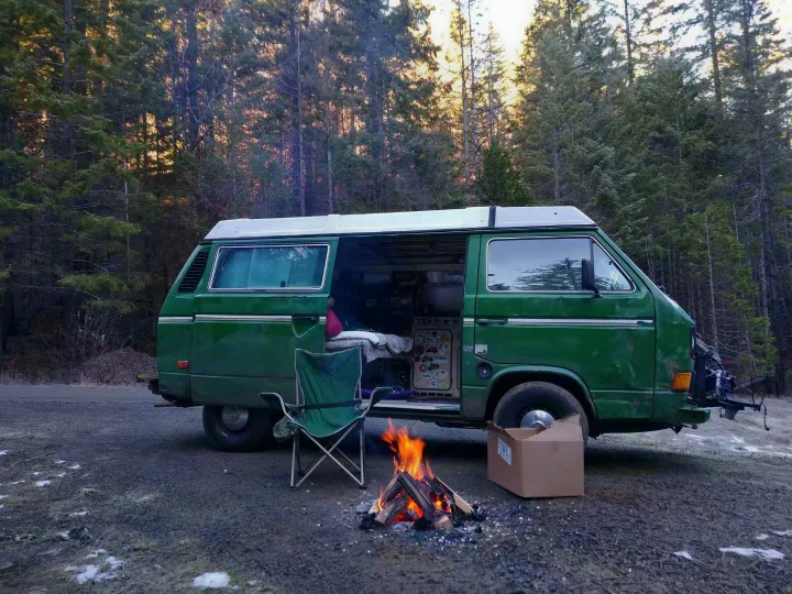 A green camper van, likely an RV rental, parked in a forest clearing with a cozy campfire and folding chair ready for camping.. Volkswagen Vanagon 1984