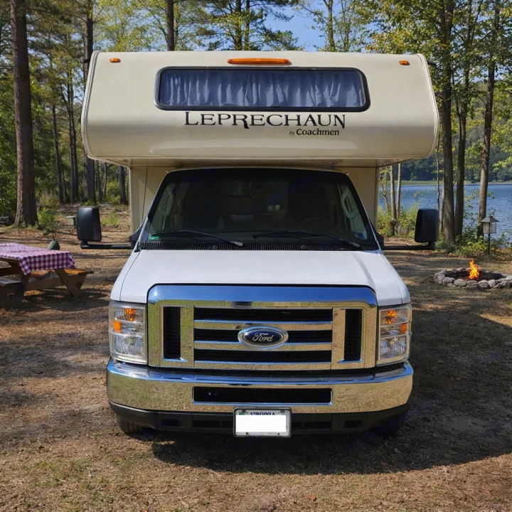 Front view of a Class C rv rental at a lakeside campsite with a picnic table and lit campfire, surrounded by trees.. Coachmen Leprechaun 2018