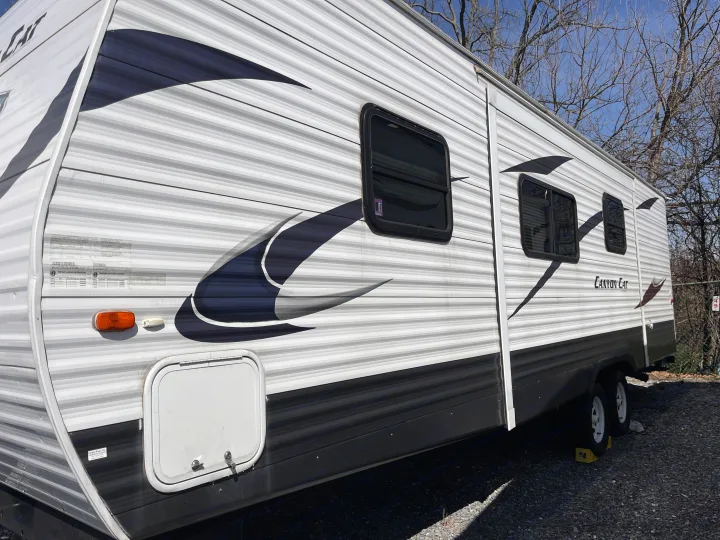 Side view of a white and grey striped "Canyon Cat" travel trailer RV rental, showing windows and an exterior storage hatch, parked on gravel.. Forest River Palomino Canyon Cat 2014