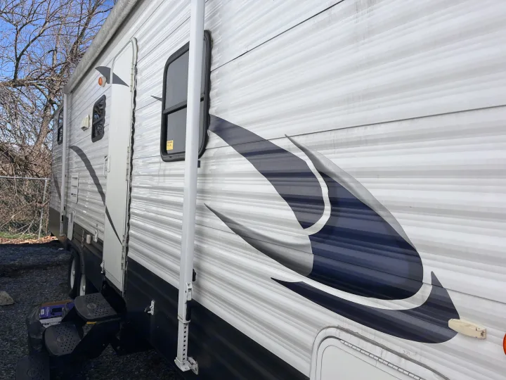 Detailed side view of an RV rental: white corrugated siding with dark graphics, entry door, windows, and steps.. Forest River Palomino Canyon Cat 2014