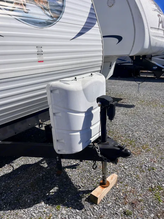 RV rental: White travel trailer's front hitch assembly, propane tank cover, and jack stand on gravel.. Forest River Palomino Canyon Cat 2014