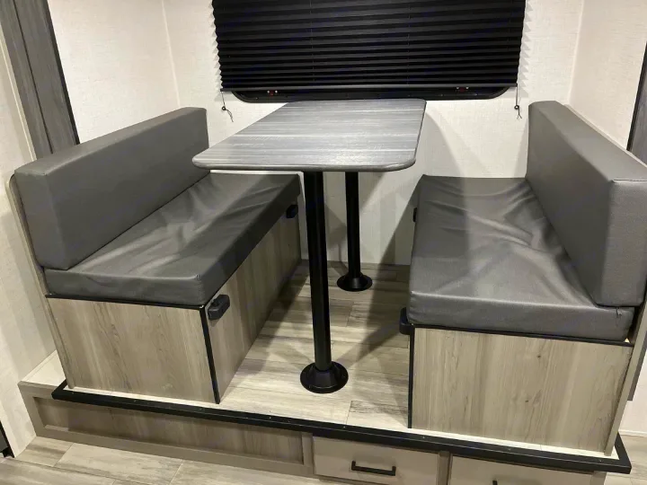Inside an RV rental: a modern dinette with gray upholstered booth seating, a light wood-grain table, and black window blind.. Keystone RV Hideout Sport 2024