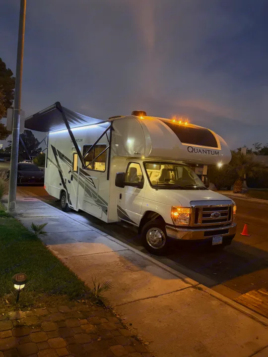 A bright white Quantum rv rental is parked at night on a street. Its awning is extended with illuminated LEDs, headlights on.. Thor Quantum 2025