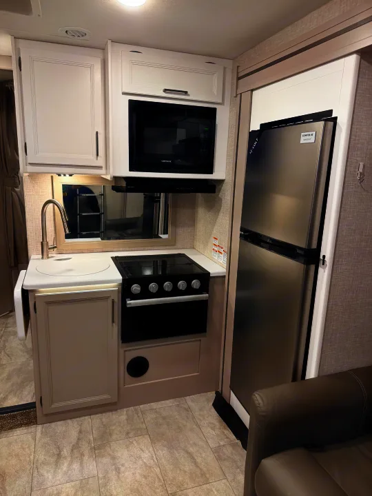 This modern RV rental kitchen features stainless steel appliances including a microwave, fridge, stovetop, oven, and sink with light cabinetry.. Thor Quantum 2025
