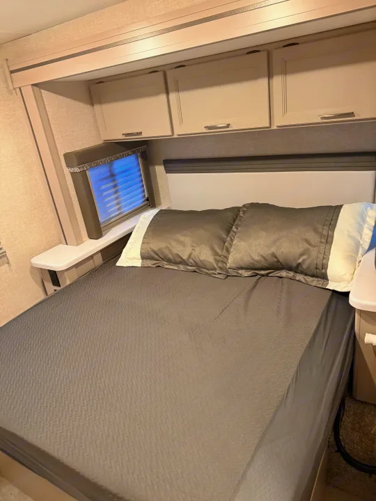 RV rental bedroom with a gray textured bedspread, padded headboard, overhead cabinets, and a window with blinds.. Thor Quantum 2025