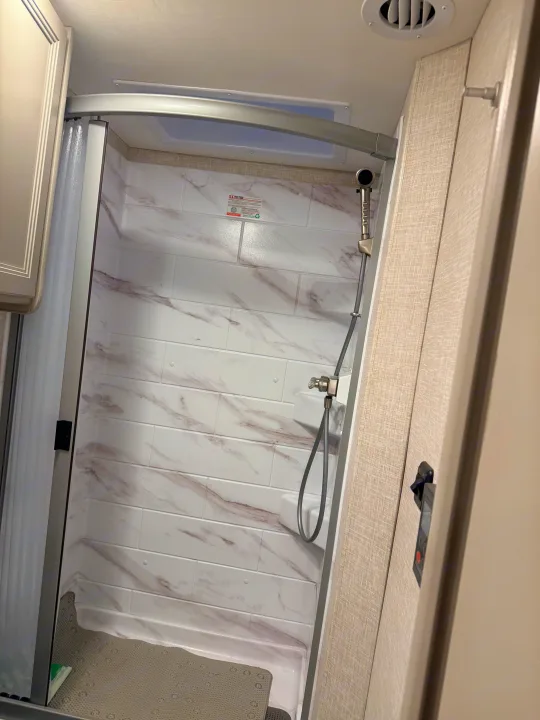RV rental shower interior with white marble-patterned tiles, a handheld showerhead, and a grey floor mat.. Thor Quantum 2025