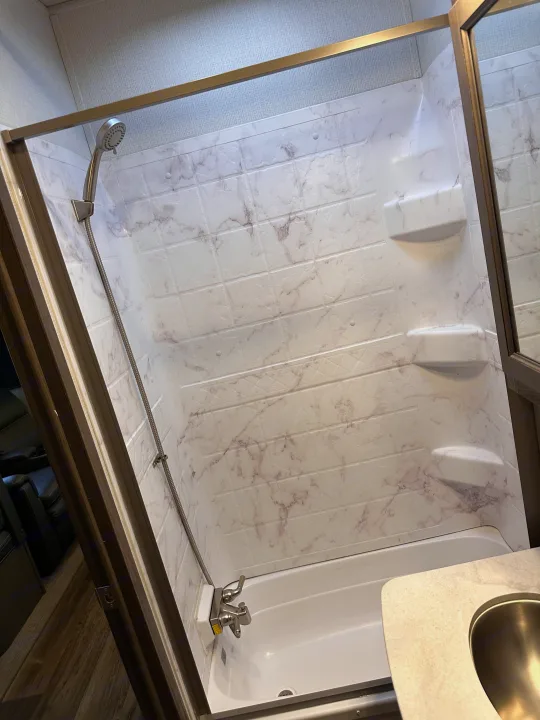 Spacious RV rental shower with a small bathtub, faux marble walls, flexible showerhead, and integrated shelves.. Keystone RV Cougar 2022
