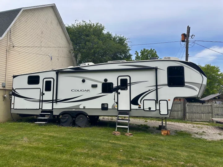 This large Cougar fifth-wheel RV rental is parked on grass and gravel next to a house with trees and a utility pole visible.. Keystone RV Cougar 2022