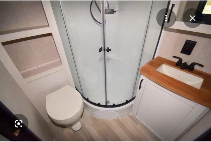This compact RV rental bathroom features a corner shower, toilet, and a modern vanity with a wood-look counter and sink.. Starcraft Autumn Ridge 2024