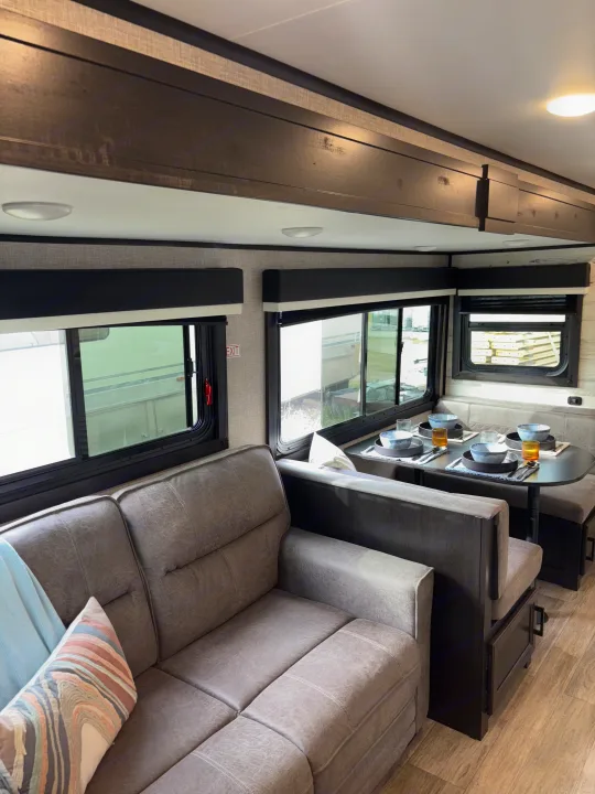 Comfortable RV rental interior with a grey sofa and a dining booth set for four, surrounded by large windows.. Jayco Jay Feather 2022