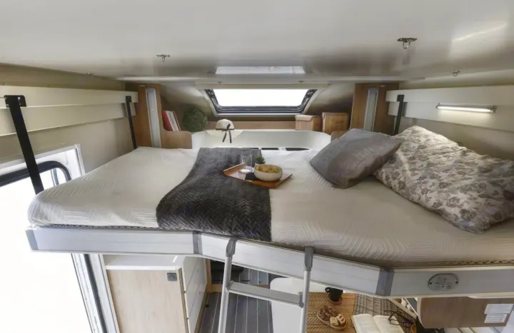 Cozy overhead sleeping area in an RV rental, complete with a made bed, breakfast tray, and ladder for access.. Ford Rollerteam 747 2026