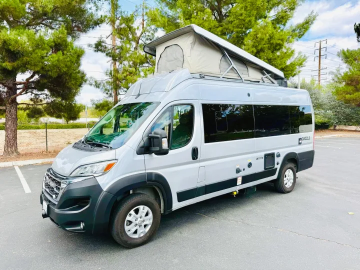A sleek silver pop-top camper van, available for rv rental, is parked outdoors. Features a raised roof tent.. Thor Dazzle 2024