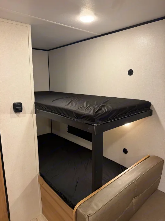 RV rental bunk beds: a compact, functional sleeping area with black mattresses, light walls, a thermostat, and a sofa.. Coleman 17B 2026