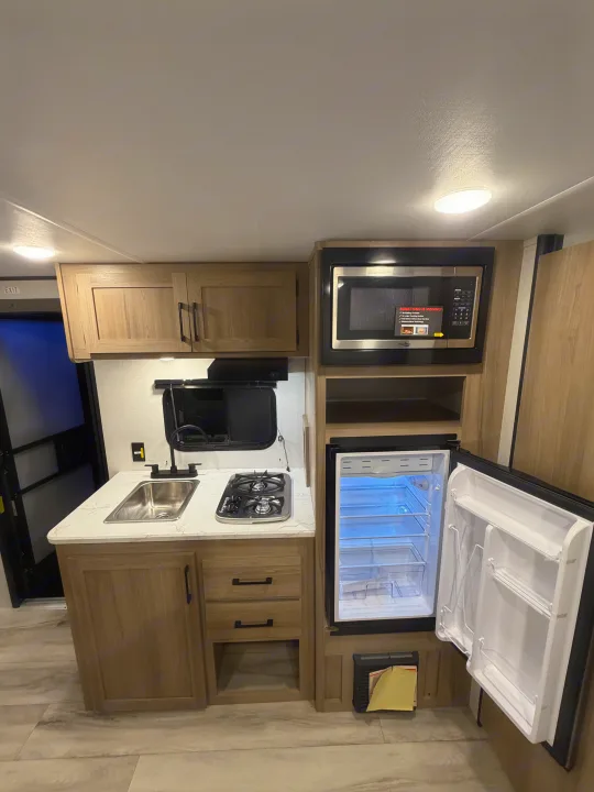 This RV rental features a modern, compact kitchen with light wood cabinets, a sink, a two-burner stovetop, microwave, and open refrigerator.. Coleman 17B 2026