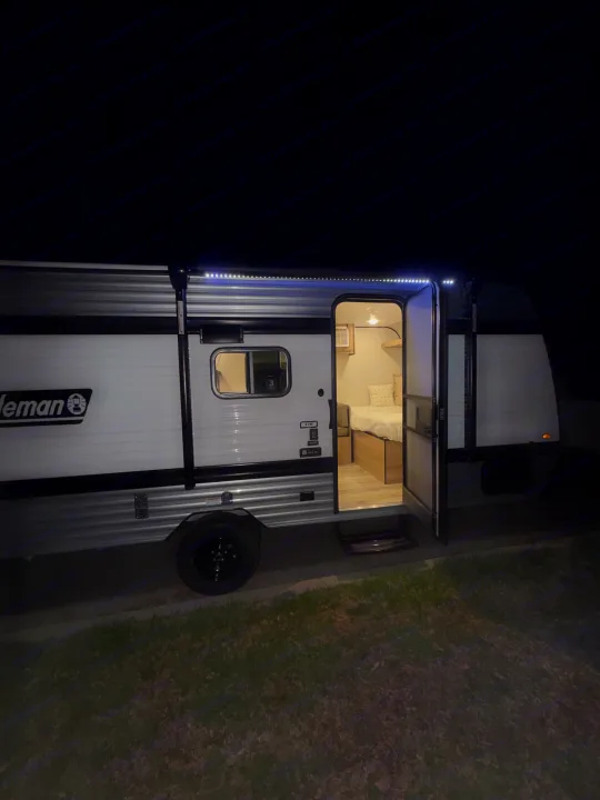 Night view of a Coleman RV rental, door open revealing a cozy bed and illuminated interior with awning lights.. Coleman 17B 2026
