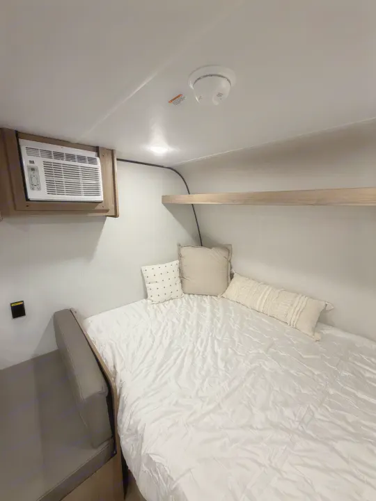 This RV rental bedroom features a comfy bed with pillows, wall-mounted AC, smoke detector, and a practical shelf.. Coleman 17B 2026