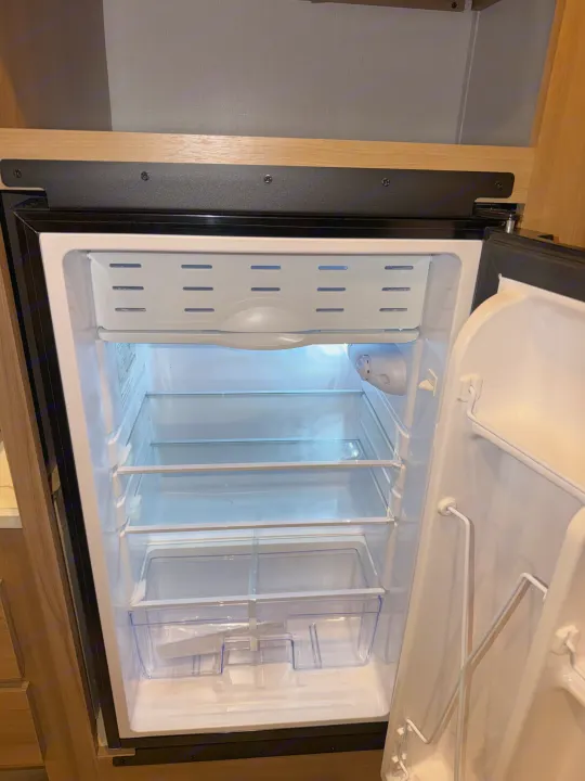 This clean, empty RV rental refrigerator features glass shelves, a top freezer, door storage, and internal lighting.. Coleman 17B 2026
