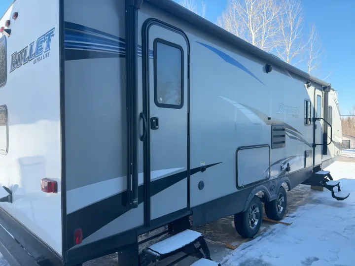 Side view of a grey Bullet Ultra Lite travel trailer RV rental, parked in snow under a blue sky, ready for adventure.. Keystone RV Bullet Ultra Lite 2019