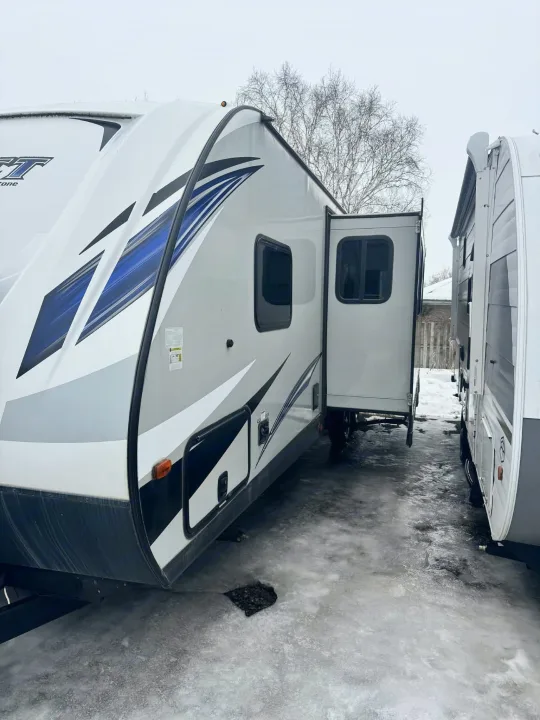 This RV rental, a white camper with blue accents, is parked with its slide-out extended on icy ground, suggesting winter conditions.. Keystone RV Bullet Ultra Lite 2019