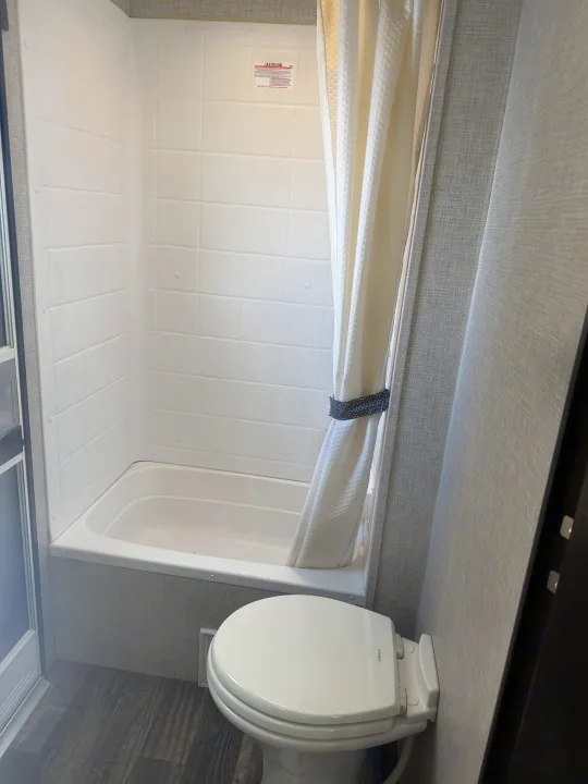 Compact RV rental bathroom features a white toilet, small tub, and shower with a textured curtain.. Keystone RV Bullet Ultra Lite 2019