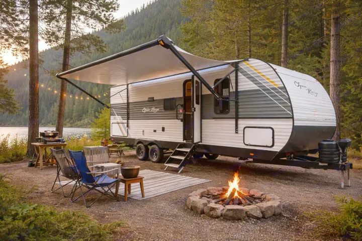 Cozy rv rental campsite: RV with awning & string lights, glowing campfire, chairs by a serene lake, surrounded by nature.. Open Range Conventional 26BHS Travel Trailer 2025
