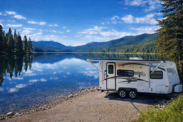 Scenic RV rental parked by a tranquil lake, reflecting a blue sky with clouds and surrounded by forested mountains.. Forest River Rockwood Mini Lite 2014