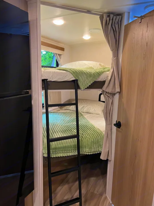 Cozy RV rental interior with bunk beds, a black ladder, soft green & white bedding, & window view. Perfect camper sleeping area.. Forest River Rockwood Mini Lite 2024