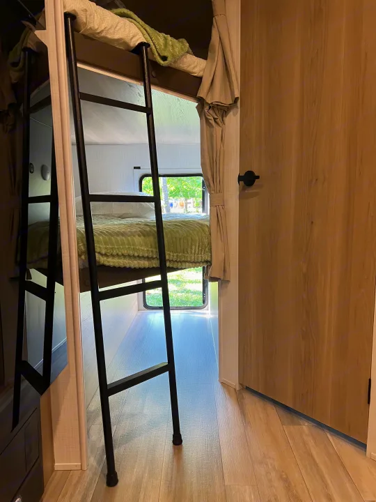 Cozy RV rental interior featuring stacked bunk beds with a ladder, green blankets, a window view of trees, and a wooden door.. Forest River Rockwood Mini Lite 2024