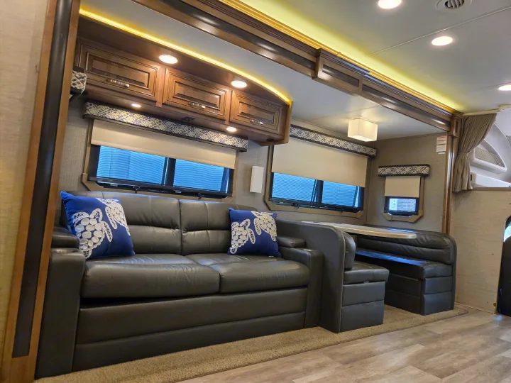 Inside an rv rental: dark leather sofa with blue pillows, adjacent dinette, overhead cabinets, and windows with blinds.. Jayco Seneca 2021