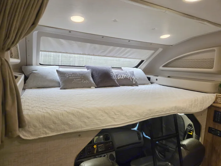 Cozy over-cab bunk bed in an RV rental, with decorative pillows, a window view, and the cockpit visible below.. Jayco Seneca 2021