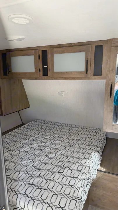 RV rental bedroom with a white and black geometric bedspread, wooden overhead cabinets, and ceiling lights.. Heartland RVs Pioneer 2021