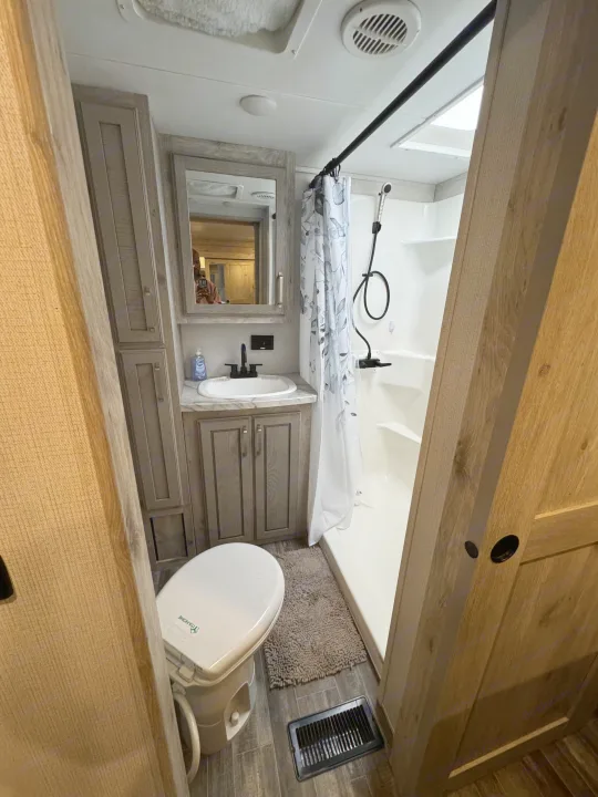Functional rv rental bathroom: toilet, vanity with sink & mirror (person visible), and shower. Modern wood-grain cabinets and floor.. Keystone RV Avalanche 2021