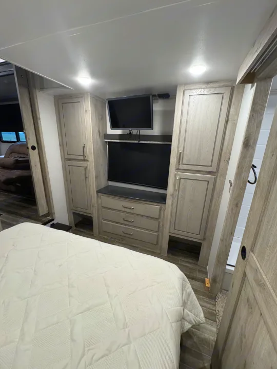 Modern RV rental bedroom featuring an entertainment center with dual TVs, dresser, storage, and a comfy white bed.. Keystone RV Avalanche 2021