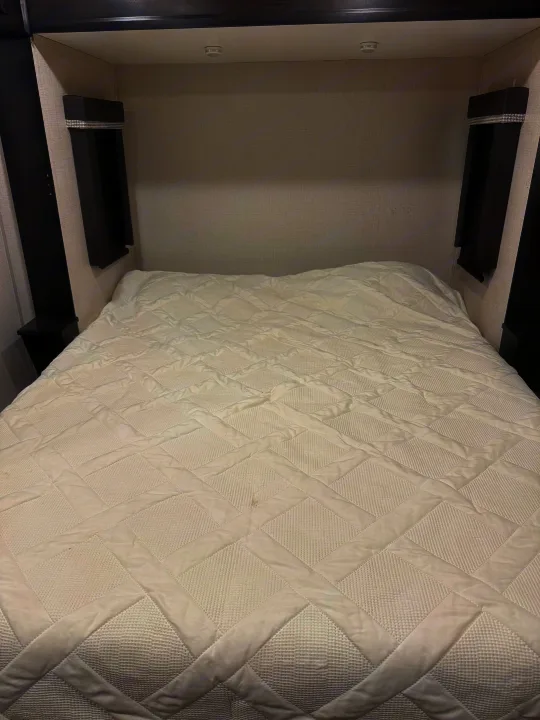 Cozy RV rental bedroom featuring a white quilted comforter on the bed, dark built-in headboard, nightstands, and overhead lights.. Keystone RV Avalanche 2021