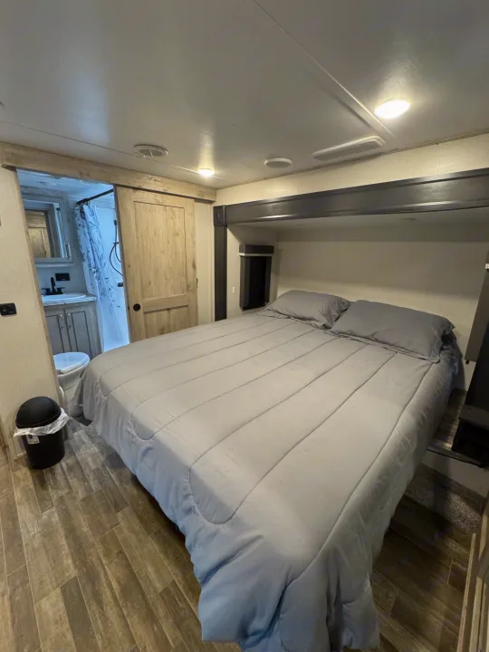 Spacious RV rental bedroom featuring a comfy grey bed, wood-look floor, and an en-suite bathroom with a sliding barn door.. Keystone RV Avalanche 2021