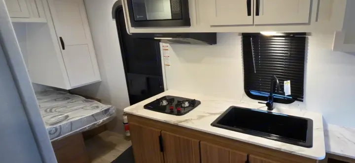 Modern RV rental interior: compact kitchen with black sink, stove, microwave, and sleek cabinets, beside a bed.. Keystone Passport 190RDWE 2026
