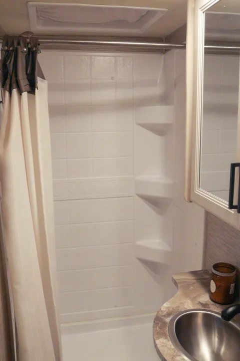 Clean RV rental bathroom showing a white shower stall with built-in shelves, a shower curtain, and a vanity with a sink and mirror.. Thor Compass 2021