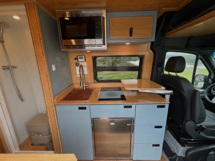 RV rental kitchen featuring microwave, sink, induction cooktop, fridge, light blue cabinets, bamboo accents. Shower and toilet visible on left.. Mercedes-Benz Sprinter 2024