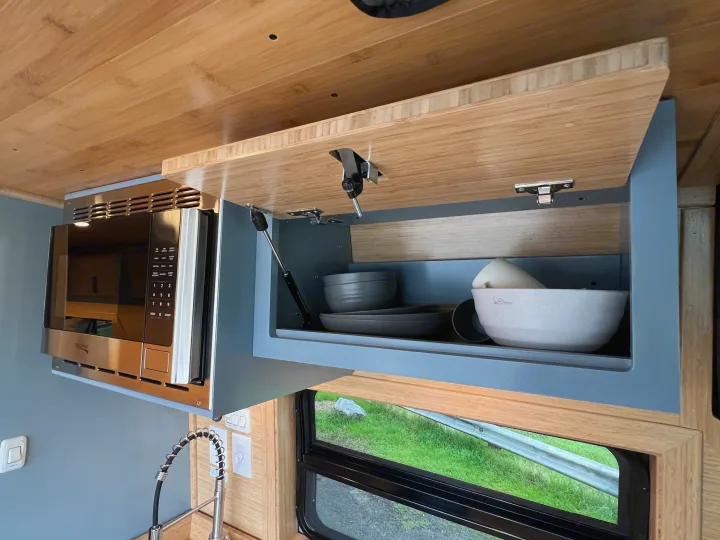 Functional RV kitchen with overhead microwave and open gray cabinet, neatly storing bowls and plates, ideal for your rv rental trip.. Mercedes-Benz Sprinter 2024