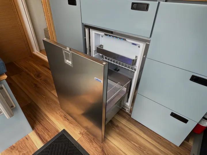 Inside an RV rental: modern kitchen with an open Isotherm fridge, light blue cabinets, wood floor, and fire extinguisher.. Mercedes-Benz Sprinter 2024