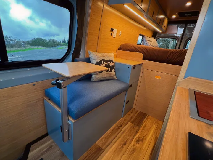 Stylish RV rental interior with a comfy bed, flexible dining table, and built-in storage amidst warm wood finishes.. Mercedes-Benz Sprinter 2024