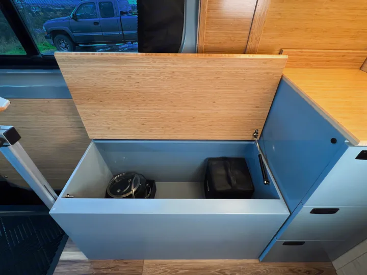 RV rental interior: an open storage compartment revealing kitchenware. Features a bamboo lid, blue cabinetry, and wood-look flooring.. Mercedes-Benz Sprinter 2024