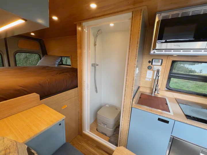RV rental interior: bed, shower with portable toilet, kitchen with sink, cooktop, and microwave. Light wood design.. Mercedes-Benz Sprinter 2024